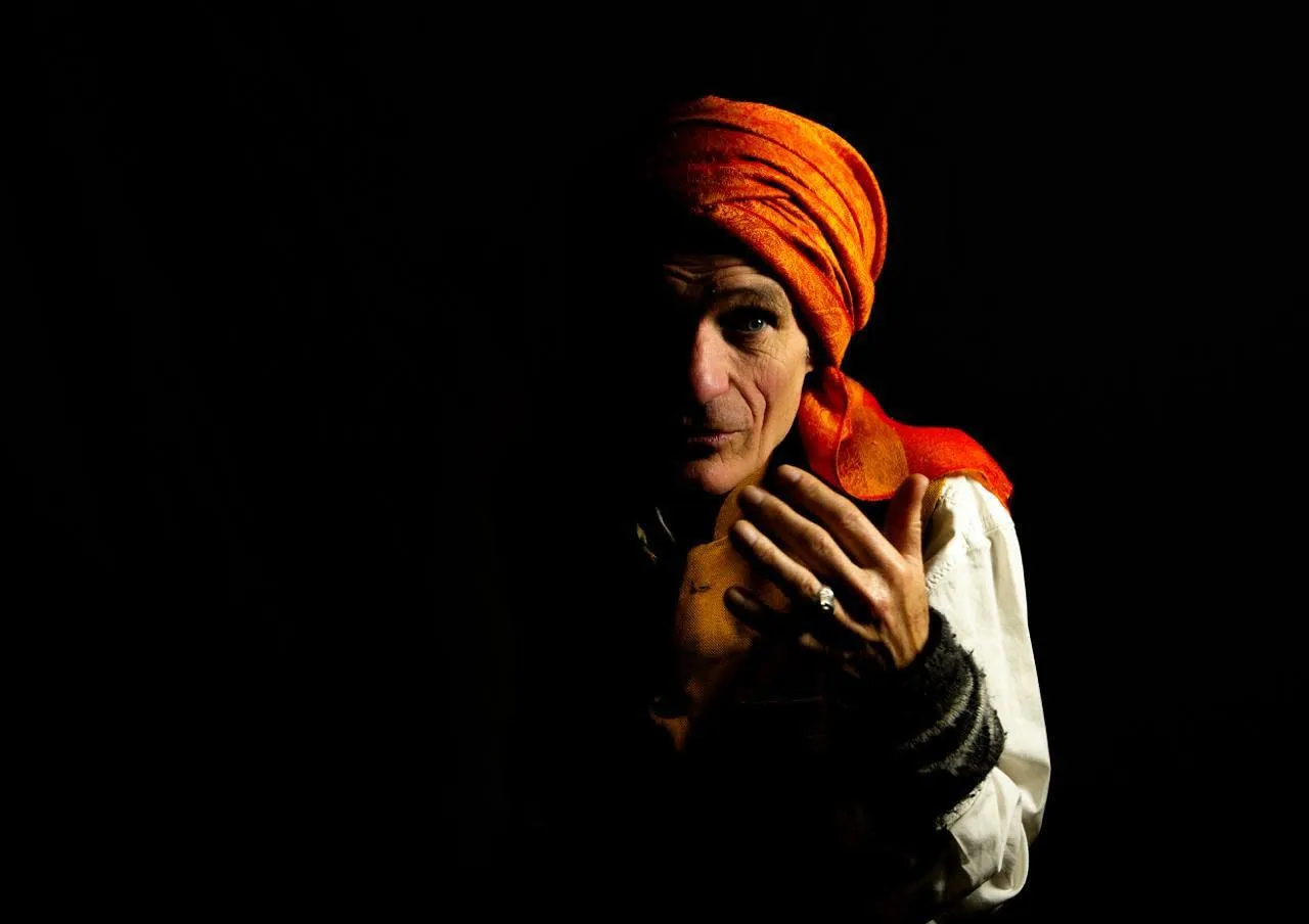 Sylvain, turban orange, surgissant de l'ombre — la main près du visage, le regard intense